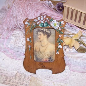 May include: A wooden picture frame with a hand-painted floral design featuring white daisies and green leaves. The frame contains a portrait of a person wearing a tiara and a double-strand necklace. The word Nice is written in cursive at the bottom.