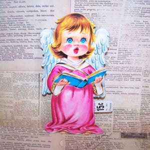 May include: A vintage illustration of a cherubic angel with blonde hair, blue eyes, and pink cheeks, holding a blue book and singing. The angel wears a pink robe and white wings, set against a backdrop of aged newspaper.