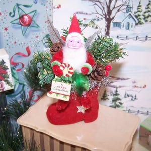 May include: A festive Christmas decoration featuring a Santa Claus figure in a red boot. The Santa has a white beard and holds a sign that reads "A Merry Christmas." The boot is adorned with a star and surrounded by greenery, pinecones, and silver tinsel.