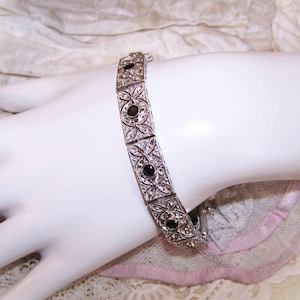 May include: Silver-toned bracelet with an ornate design featuring clear and black stones. The bracelet is composed of rectangular links with floral and geometric patterns. The bracelet is displayed on a white surface.