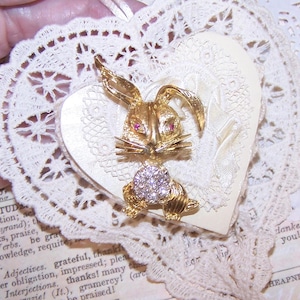 May include: A gold-toned bunny brooch with red gem eyes and a crystal-studded belly, set on a cream-colored heart-shaped card. The card is adorned with delicate lace and a vintage aesthetic.