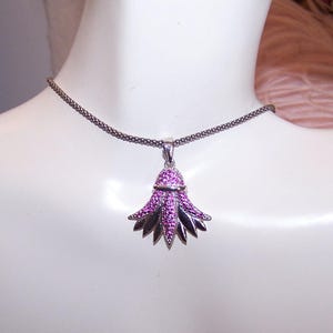 May include: A silver-toned necklace with a pendant shaped like a stylized flower. The pendant features a cluster of small, pink gemstones and dark silver-colored petals. The necklace is displayed on a white mannequin.
