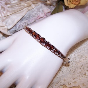 May include: A silver bracelet with a row of oval, deep red gemstones. The bracelet is set on a white surface, showcasing the gemstones' color and the silver's shine. The clasp is visible.