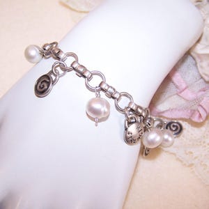 May include: A silver charm bracelet featuring multiple charms, including pearls and spiral designs. The bracelet is composed of interconnected links and is adorned with several dangling charms. The pearls are white and the metal has a slightly aged appearance.
