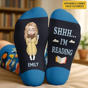 Op de afbeelding: Een paar marineblauwe sokken met blauwe tenen. Eén sok toont een cartoon van een lezende persoon met de naam "EMILY". De andere sok zegt "SHHH... I'M READING" met een boekafbeelding. De tekst "APPEARANCE & NAMES CAN BE CUSTOMIZED" staat rechtsboven.