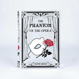 May include: A clear, rectangular book-shaped object featuring the title "The Phantom of the Opera" in black text. The cover design includes a white mask, a red rose, and decorative black borders. The author's name, Gaston Leroux, is also displayed.