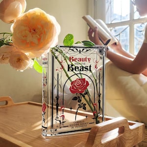 K&ouml;nnte beinhalten: Eine durchsichtige Glasvase in Form eines Buches mit dem Titel "Die Sch&ouml;ne und das Biest" auf der Vorderseite. Die Vase ist mit wei&szlig;en Rosen und gr&uuml;nen Stielen gef&uuml;llt. Die Vase steht auf einem Holztisch.