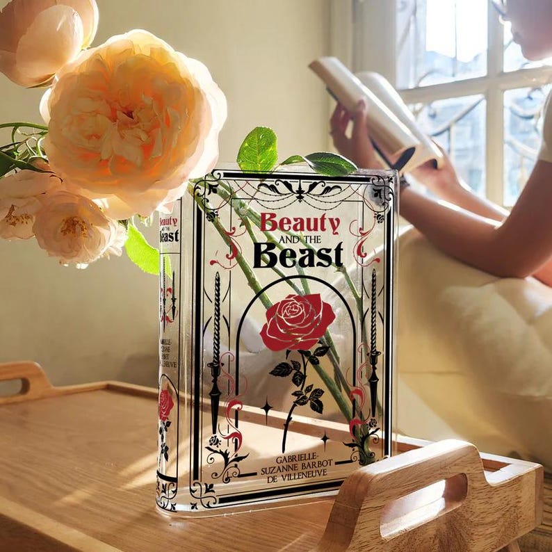 K&ouml;nnte beinhalten: Eine durchsichtige Glasvase in Form eines Buches mit dem Titel "Die Sch&ouml;ne und das Biest" auf der Vorderseite. Die Vase ist mit wei&szlig;en Rosen und gr&uuml;nen Stielen gef&uuml;llt. Die Vase steht auf einem Holztisch.