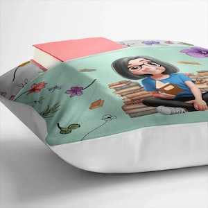 May include: A cushion with a light teal illustration of a person reading, surrounded by books and flowers. A pink book rests on top. The cushion has a white border. The design includes illustrations of books and flowers.