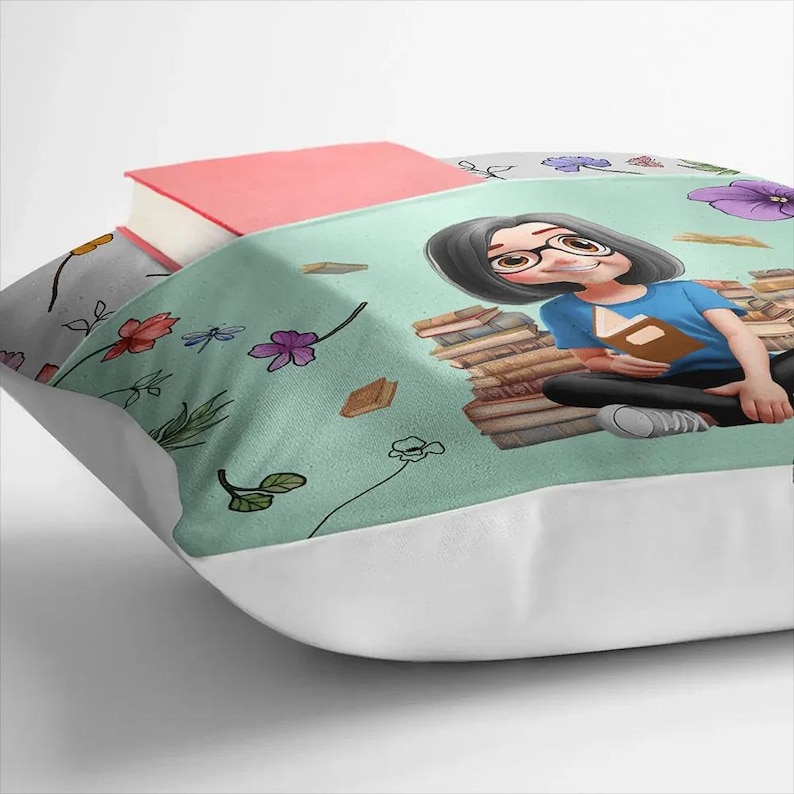 May include: A cushion with a light teal illustration of a person reading, surrounded by books and flowers. A pink book rests on top. The cushion has a white border. The design includes illustrations of books and flowers.