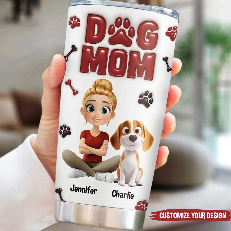 Op de afbeelding: Een witte roestvrijstalen beker met een rood "Dog Mom"-ontwerp. De beker heeft een cartoonillustratie van een vrouw met blond haar en een hond. De tekst "Jennifer" en "Charlie" staat onder de illustratie.