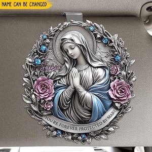 Könnte beinhalten: Ein silberfarbener Auto-Anhänger mit einer Darstellung der Jungfrau Maria in einem blauen Gewand und einer Blumenkrone. Der Anhänger trägt den Text "You're forever protected by Mary" und den Namen "Sophia".