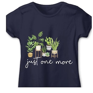 Puede incluir: Camiseta azul marino con una ilustraci&oacute;n de acuarela de plantas en macetas, incluyendo una monstera y cactus. El dise&ntilde;o incluye el texto "just one more" en cursiva debajo de las plantas. Cuello redondo cl&aacute;sico.