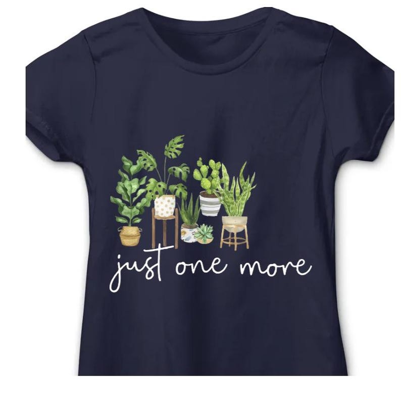 Puede incluir: Camiseta azul marino con una ilustraci&oacute;n de acuarela de plantas en macetas, incluyendo una monstera y cactus. El dise&ntilde;o incluye el texto "just one more" en cursiva debajo de las plantas. Cuello redondo cl&aacute;sico.