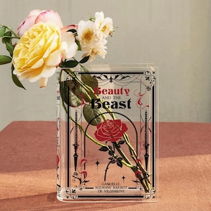 K&ouml;nnte beinhalten: Eine durchsichtige Acryl-Vase in Form eines Buches mit dem Titel "Die Sch&ouml;ne und das Biest" auf der Vorderseite. Die Vase enth&auml;lt eine rote Rose und schwarze und goldene Verzierungen.