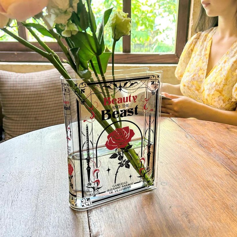K&ouml;nnte beinhalten: Eine durchsichtige Glasvase in Form eines Buches mit einer schwarz-wei&szlig;en Illustration einer Rose und dem Text "Die Sch&ouml;ne und das Biest". Die Vase ist mit Wasser und einer einzelnen roten Rose gef&uuml;llt.