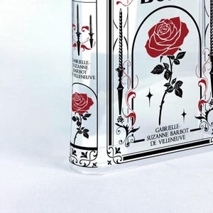 K&ouml;nnte beinhalten: Ein klares Glasbuch mit einem schwarz-wei&szlig;en Blumendesign und einer roten Rose. Der Text auf dem Buch lautet "Gabrielle-Suzanne Barbot de Villeneuve".
