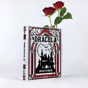 Puede incluir: Un jarrón de vidrio transparente con forma de libro con una cubierta roja y negra. La cubierta presenta el título "Drácula" y el autor "Bram Stoker". El jarrón está lleno de rosas rojas.