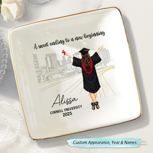 May include: A white ceramic trinket tray with a gold rim features a black and white illustration of a city skyline and a graduate walking away from the viewer. The text "A sweet ending to a new beginning" is at the top of the tray. The text "Alissa, Cornell University, 2025" is at the bottom of the tray.