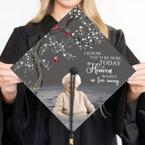 Puede incluir: Un gorro de graduación con una foto de una mujer mayor y el texto "Sé que estarías aquí hoy si el cielo no estuviera tan lejos". El gorro está decorado con un árbol con cardenales rojos y copos de nieve blancos.