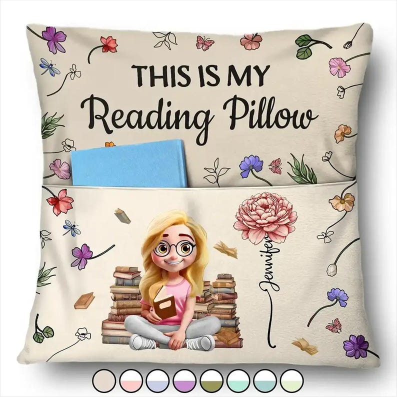May include: Cream reading pillow with the text "THIS IS MY Reading Pillow." Features a pocket with a blue book, a cartoon illustration of a girl with glasses, and a stack of books. Decorated with floral and butterfly accents.