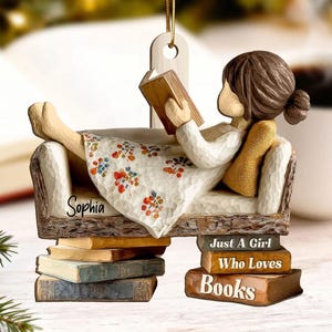 May include: A decorative ornament depicting a girl reading a book, reclining on a carved sofa. The sofa is supported by a stack of books. The girl wears a white dress with a floral pattern. The ornament has the text "Sophia" and "Just A Girl Who Loves Books".