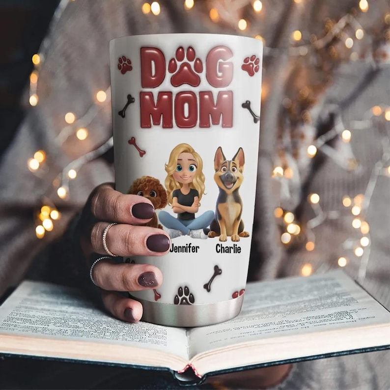 Op de afbeelding: Een witte roestvrijstalen tumbler met een rood "Dog Mom"-ontwerp. De tumbler heeft een cartoonafbeelding van een vrouw met twee honden, een bruine en een zwart-bruine. De tekst "Jennifer" en "Charlie" staat onder de honden.