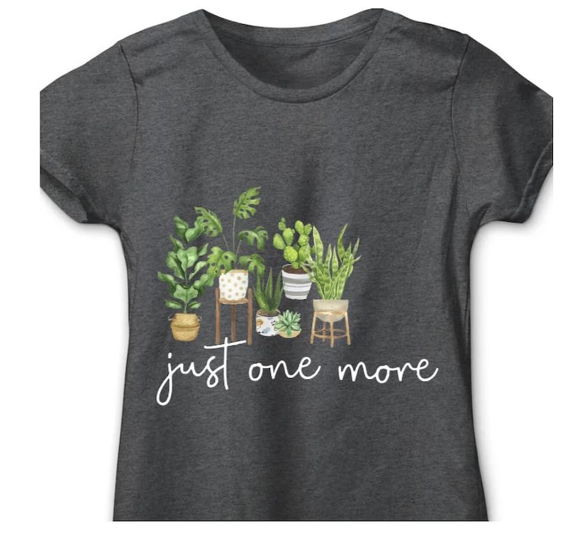 Puede incluir: Camiseta gris oscuro con una ilustraci&oacute;n de acuarela de varias plantas en macetas, incluyendo una monstera, un cactus y una sansevieria. La frase "just one more" est&aacute; escrita en cursiva debajo de las plantas.