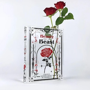 Può includere: Un vaso di vetro trasparente a forma di libro con un design in bianco e nero. Il vaso presenta il titolo "La Bella e la Bestia" e un rosa rossa all'interno.
