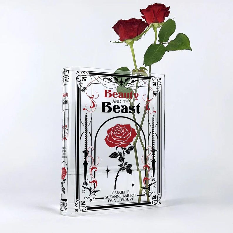 K&ouml;nnte beinhalten: Eine durchsichtige Glasvase in Form eines Buches mit einem schwarz-wei&szlig;en Design. Die Vase tr&auml;gt den Titel "Die Sch&ouml;ne und das Biest" und eine rote Rose im Inneren.
