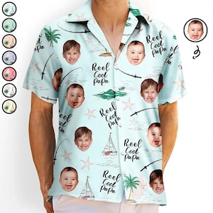 May include: Light blue short-sleeve button-down shirt with a tropical design. The shirt features images of babies, sailboats, fishing rods, palm trees, and the text "Reel Cool Papa".