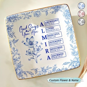 Puede incluir: Plato cuadrado de cerámica con diseño floral azul y borde dorado. El plato presenta el texto "Dios dice que eres" y una lista de palabras con versículos bíblicos. Las palabras son "Abundante", "Amado", "Misericordioso", "Destinado", "Restaurado" y "Adornado".