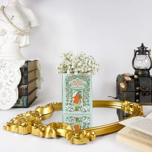 Könnte beinhalten: Eine Vase aus einem Buch mit dem Titel "The Secret Garden" enthält weiße Blumen. Die Vase steht auf einem goldfarbenen, verzierten, ovalen Spiegel. Eine antike Laterne und ein Stapel Bücher befinden sich im Hintergrund.