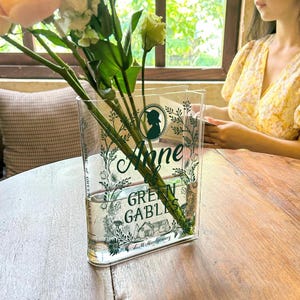 May include: A clear glass vase shaped like a book with the title "Anne of Green Gables" printed on it. The vase is filled with water and flowers.
