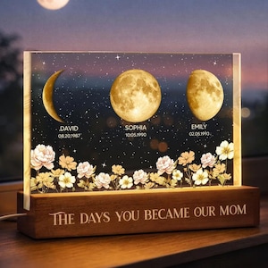 May include: A personalized night light with three moon phases, names, and dates, set against a starry sky background. The base reads "THE DAYS YOU BECAME OUR MOM". Floral accents adorn the bottom. The light is on a wooden base.