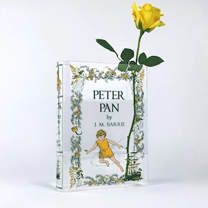 Può includere: Un vaso di vetro trasparente a forma di libro, con il titolo "Peter Pan" di J.M. Barrie. Il vaso presenta illustrazioni di Peter Pan, fate e una nave. Una rosa gialla è posta all'interno del vaso.