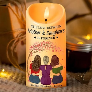 Puede incluir: Una vela decorativa con un brillo cálido, con el texto "THE LOVE BETWEEN Mother & Daughters IS FOREVER". El diseño de la vela incluye una ilustración de tres figuras bajo un árbol en flor, con los nombres Jasmine, Elizabeth y Jessica.