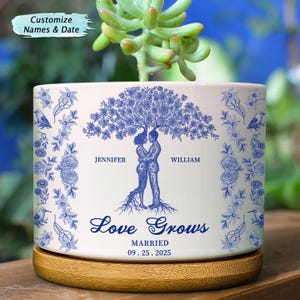 Könnte beinhalten: Weißer Keramik-Pflanztopf mit blauem Baumdesign, das ein sich umarmendes Paar zeigt. Der Topf trägt die Worte "Love Grows" und das Datum "09.25.2025". Der Topf steht auf einem Holzsockel. Der Text "Customize Names & Date" steht oben.