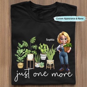 Puede incluir: Camiseta negra con una ilustraci&oacute;n de dibujos animados de una mujer con plantas y el texto "just one more". La mujer sostiene una cesta de plantas. La camiseta tambi&eacute;n tiene el nombre "Sophia".