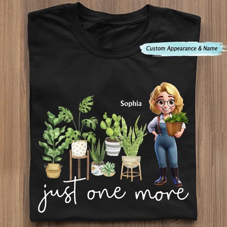Puede incluir: Camiseta negra con una ilustraci&oacute;n de dibujos animados de una mujer con plantas y el texto "just one more". La mujer sostiene una cesta de plantas. La camiseta tambi&eacute;n tiene el nombre "Sophia".
