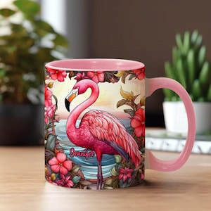 Op de afbeelding: Roze keramische mok met een flamingo-illustratie met de naam "Jennifer". De mok heeft een roze binnenkant en handvat en is versierd met bloemen en een strandscène. De mok staat op een houten oppervlak.