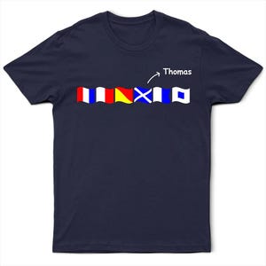 Könnte beinhalten: Marineblaues T-Shirt mit einer Reihe farbenfroher nautischer Signalflaggen. Die Flaggen sind rot, weiß, blau und gelb. Der Name "Thomas" steht in weißer Schrift über den Flaggen.