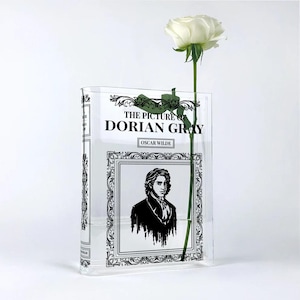 Może przedstawiać: Przezroczysty wazon akrylowy w kształcie książki, z tytułem "THE PICTURE OF DORIAN GRAY" i portretem Oscara Wilde'a. Biała róża z długą zieloną łodygą umieszczona jest w wazonie na białym tle.