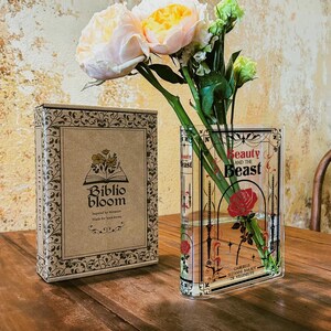K&ouml;nnte beinhalten: Eine durchsichtige Glasvase in Form eines Buches mit dem Titel "Die Sch&ouml;ne und das Biest" auf der Vorderseite. Die Vase ist mit rosa und wei&szlig;en Blumen und einer einzelnen roten Rose gef&uuml;llt.