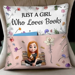 May include: A cushion featuring the text "JUST A GIRL Who Loves Books" and a cartoon illustration of a girl with red hair sitting in front of a stack of books. The cushion has a pocket and is decorated with flowers.