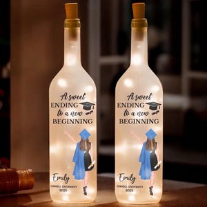 Op de afbeelding: Twee verlichte glazen flessen met kurken. Elke fles heeft de tekst "A sweet ENDING to a new BEGINNING" en een afstudeerillustratie. De naam "Emily" en "CORNELL UNIVERSITY 2026" staan ook op de flessen.