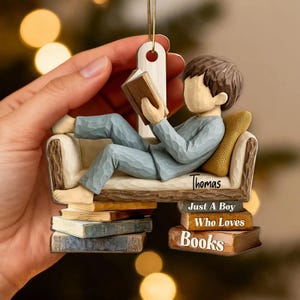 May include: A decorative ornament featuring a boy reading a book while reclining on a miniature sofa. The ornament is light blue and brown, with the name "Thomas" and the text "Just A Boy Who Loves Books".