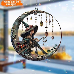May include: A green and gold moon-shaped hanging ornament with a witch sitting on it. The witch is wearing a green dress and a pointed hat with a red flower. The ornament has a name, "Jennifer", written on it.