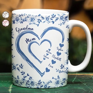 May include: White ceramic mug with a blue floral pattern and a heart-shaped design. The mug features the words "Grandma" and "Mom", plus names like Sophia and Elizabeth. The mug has a white handle and is decorated with blue flowers.