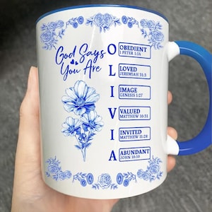 Puede incluir: Taza de cerámica blanca con interior y asa azules. La taza presenta el texto "God Says You Are" y la palabra "OLIVIA" con afirmaciones positivas y versículos bíblicos. Diseños florales azules decoran la taza.